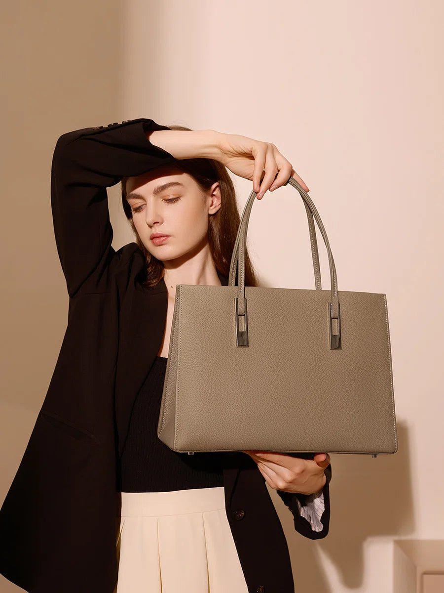 Model lifting a taupe leather tote by its top handles, showing the pebbled texture and structured silhouette