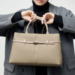 Model holding Cuirae taupe pebbled genuine leather handbag by top handles, front view showing belt buckle detail