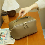 Cuirae taupe pebbled genuine leather makeup bag with top handle, hand carry on wooden table with coffee cup and newspaper