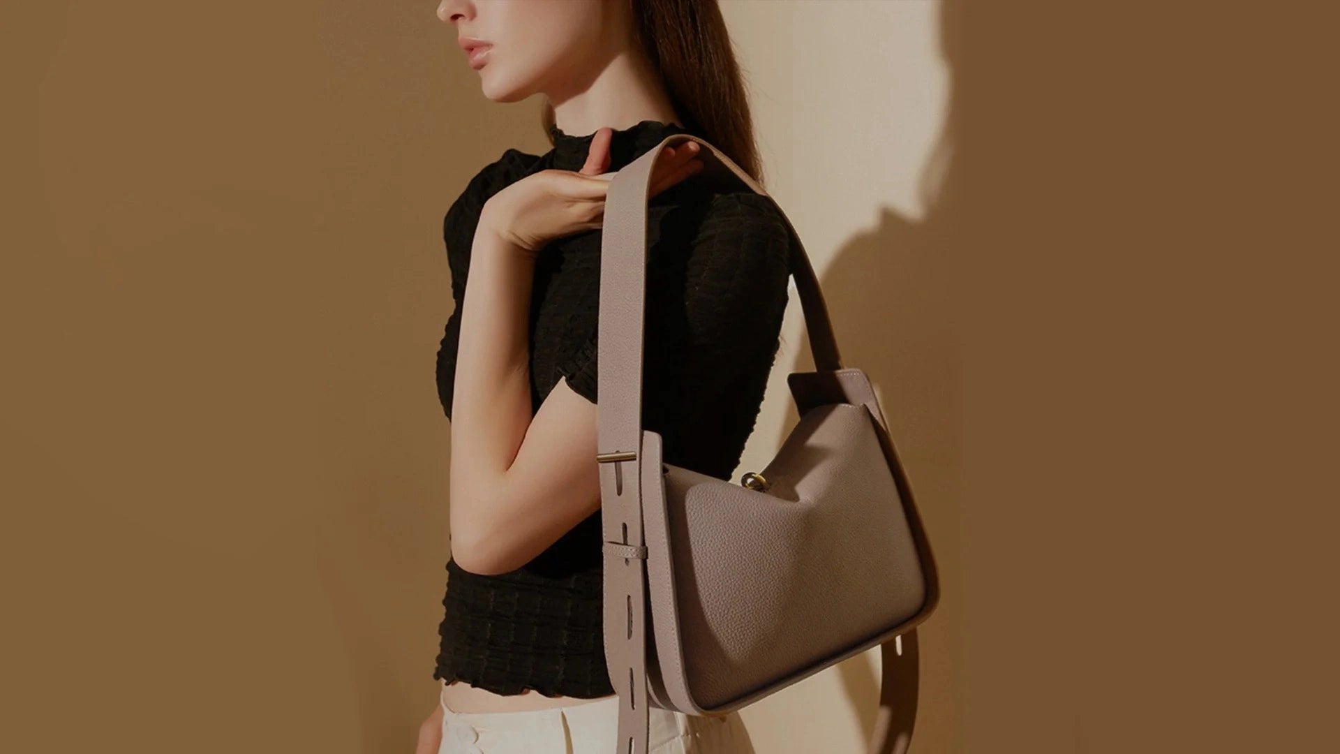 A woman in a black top carrying a taupe Cuirae 100% real leather shoulder bag. The wide strap and pebbled leather are built to last.