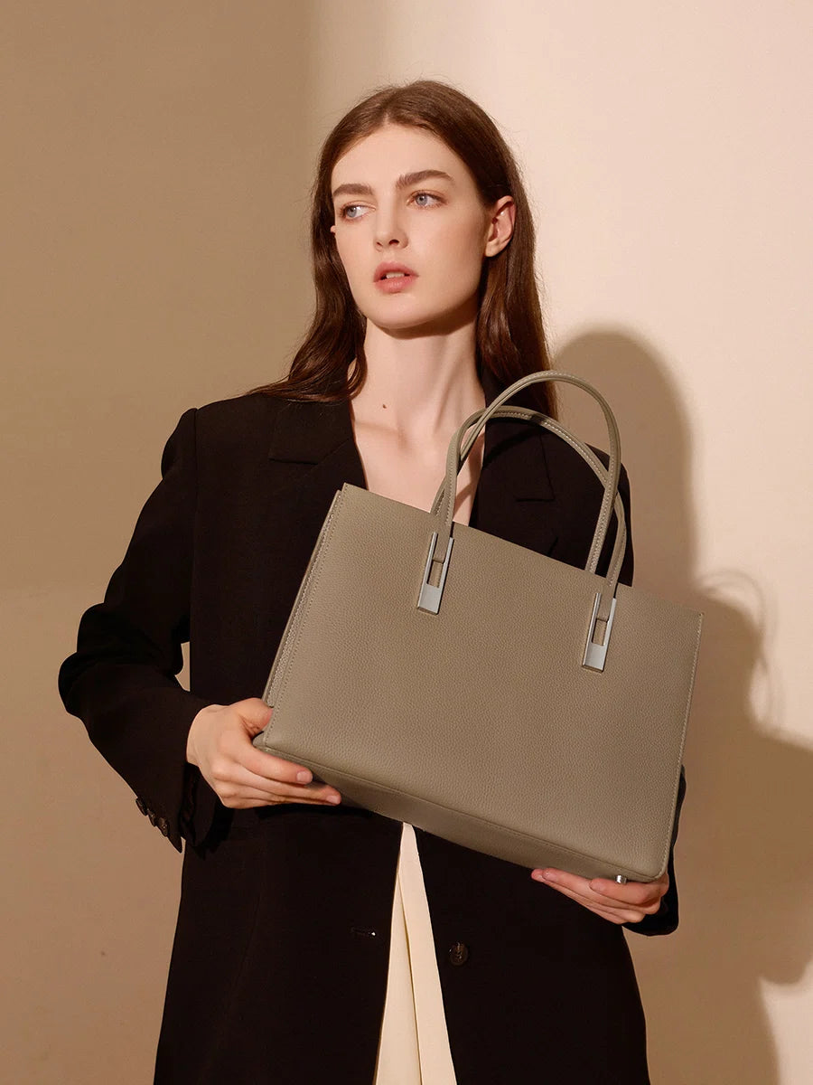 Model holding a taupe structured leather tote in front of her, showing its rectangular shape and silver-tone accents