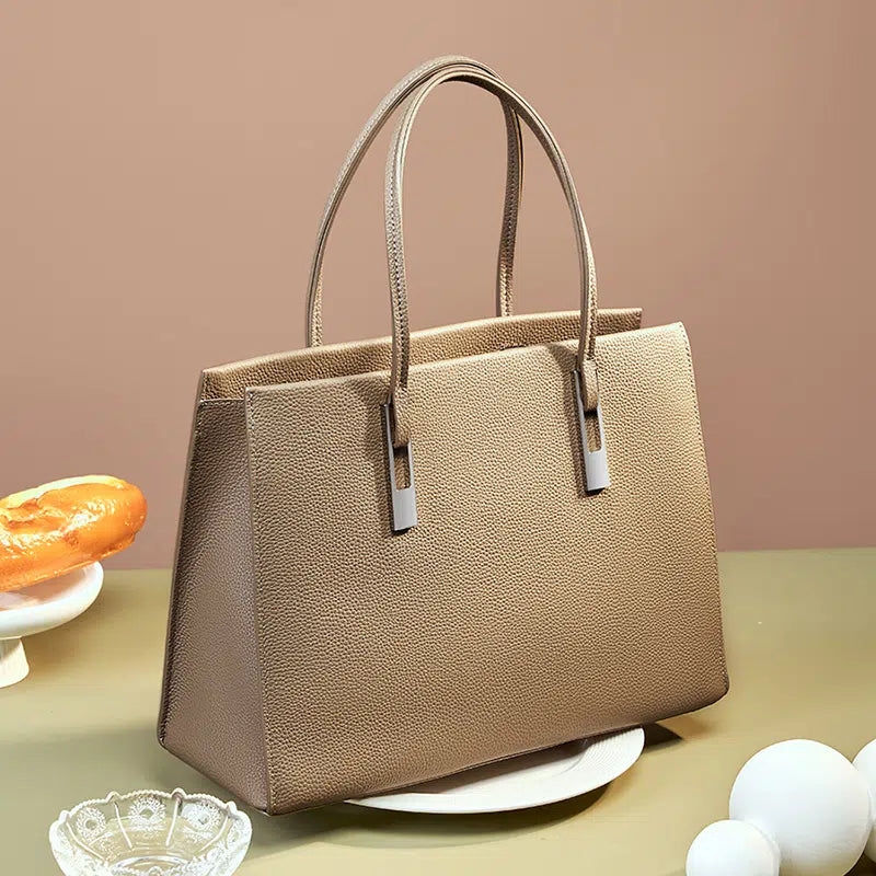 Taupe structured leather tote with pebbled texture, twin handles, and silver-tone accents displayed on a tabletop.
