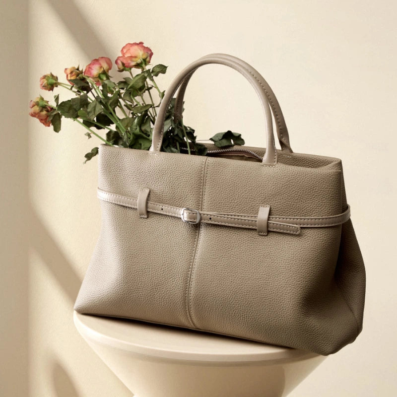A Cuirae taupe top grain cowhide handbag filled with pink roses resting on a pedestal.