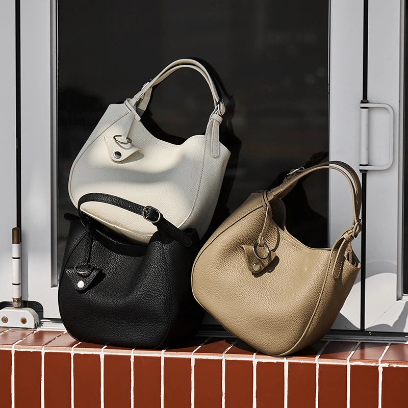 Three Cuirae top grain cowhide bucket bags in white, black, and taupe resting on a window sill in the sun.