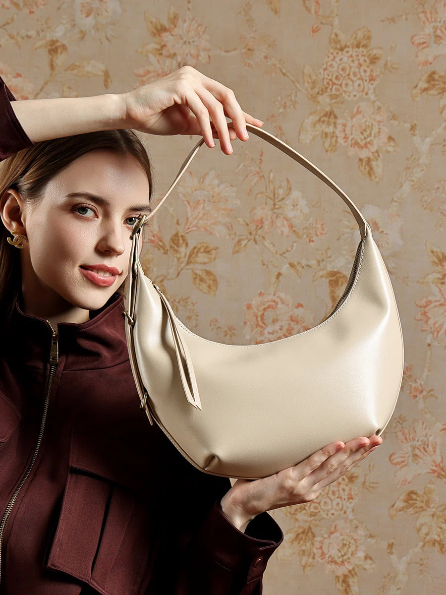 A woman in a dark red outfit holding up the Cuirae white genuine leather handbag, standing in front of floral wallpaper.