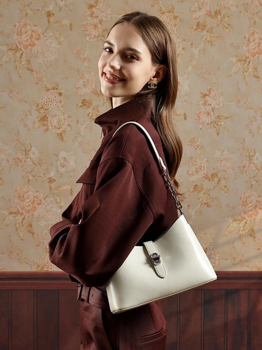 A woman in a dark red outfit carrying the Cuirae white genuine leather handbag on her shoulder, standing in front of floral wallpaper.