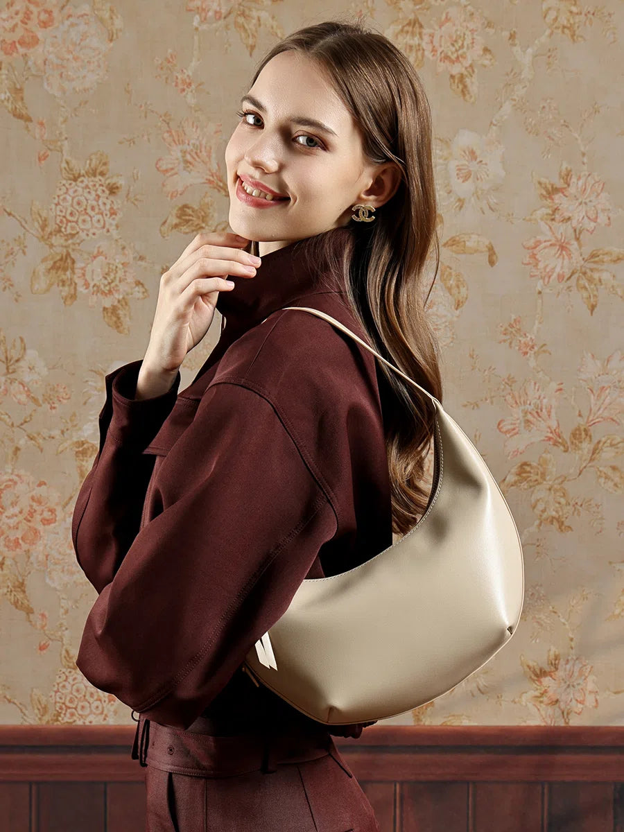 A smiling woman in a dark red outfit wearing the Cuirae white top grain cowhide handbag on her shoulder, standing in front of floral wallpaper.