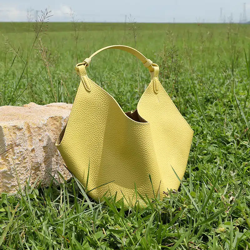 A Cuirae yellow top grain cowhide bucket bag resting in the green grass next to a stone.