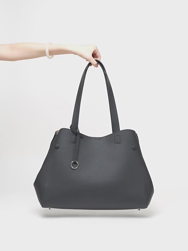 Black leather work tote shown by hand, highlighting its shape, shoulder handles, and ring accent.