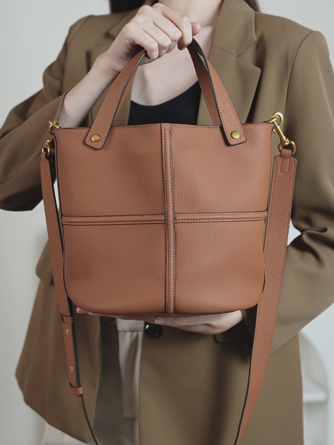 Model holding a brown pebbled leather bucket bag, showing its rounded shape, raised cross seams, and gold-tone handle rivets.
